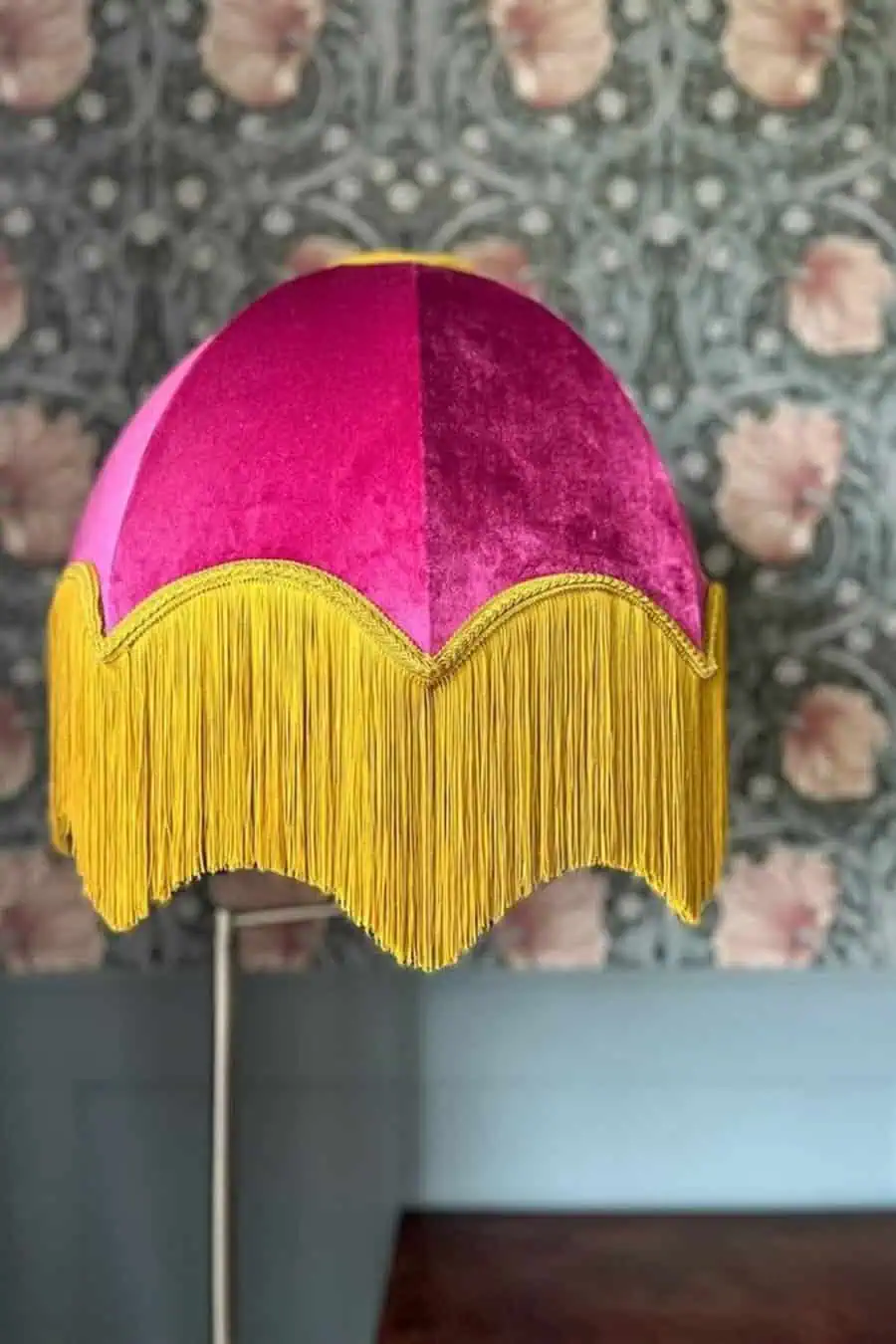 Eclectic pink velvet lampshade with yellow fringe detail and floral wallpaper background.