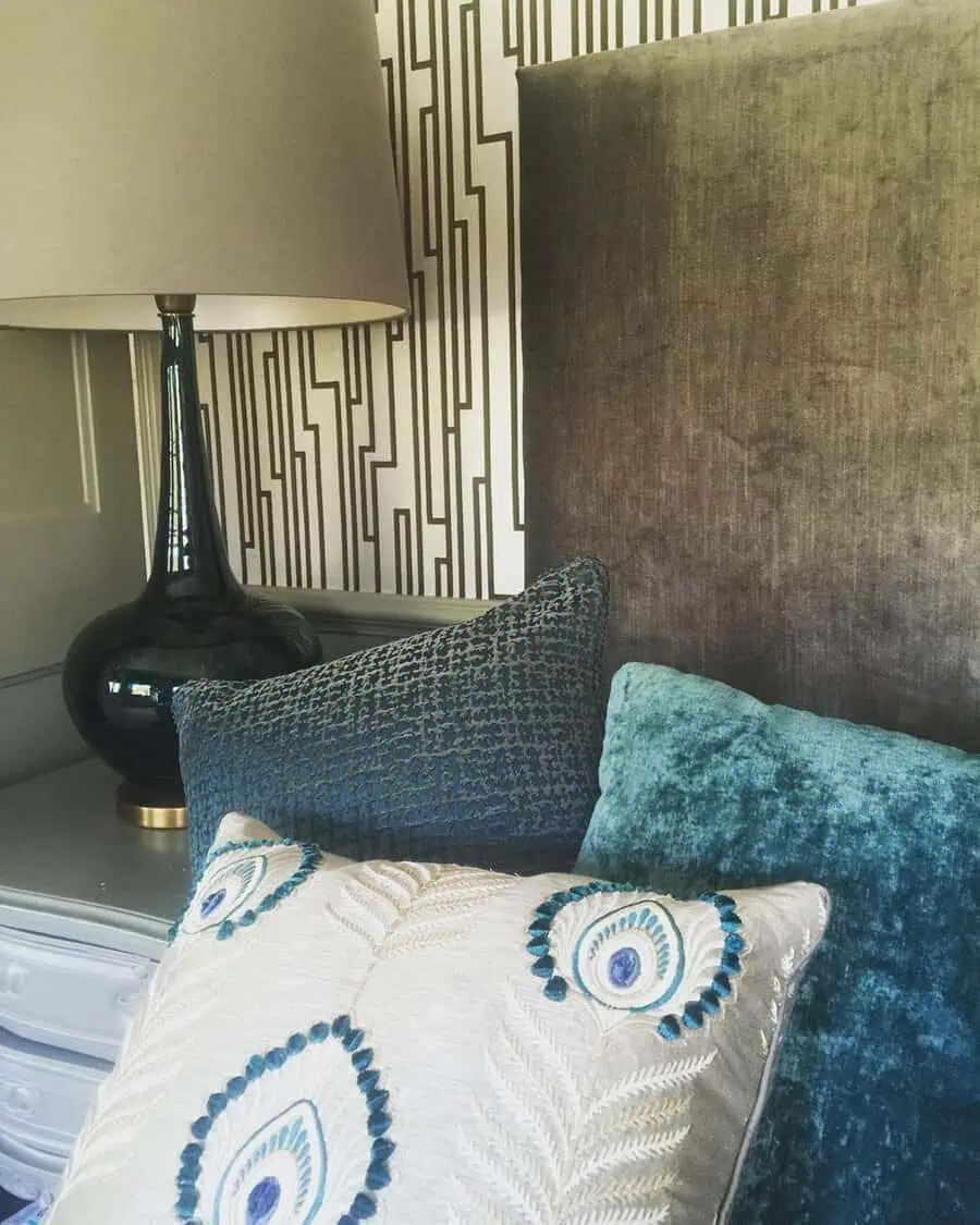 Bedroom with taupe headboard, teal pillows, peacock-inspired cushion, and a modern geometric lamp.