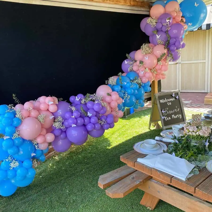 Colorful balloon arch with pink, purple, and blue balloons at an outdoor tea party setup.