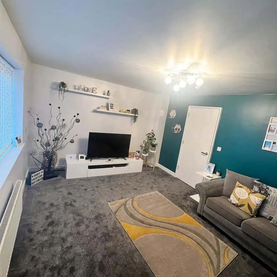 A modern living room featuring a crisp white and teal accent wall, gray carpeting, and cozy seating with geometric-patterned cushions.