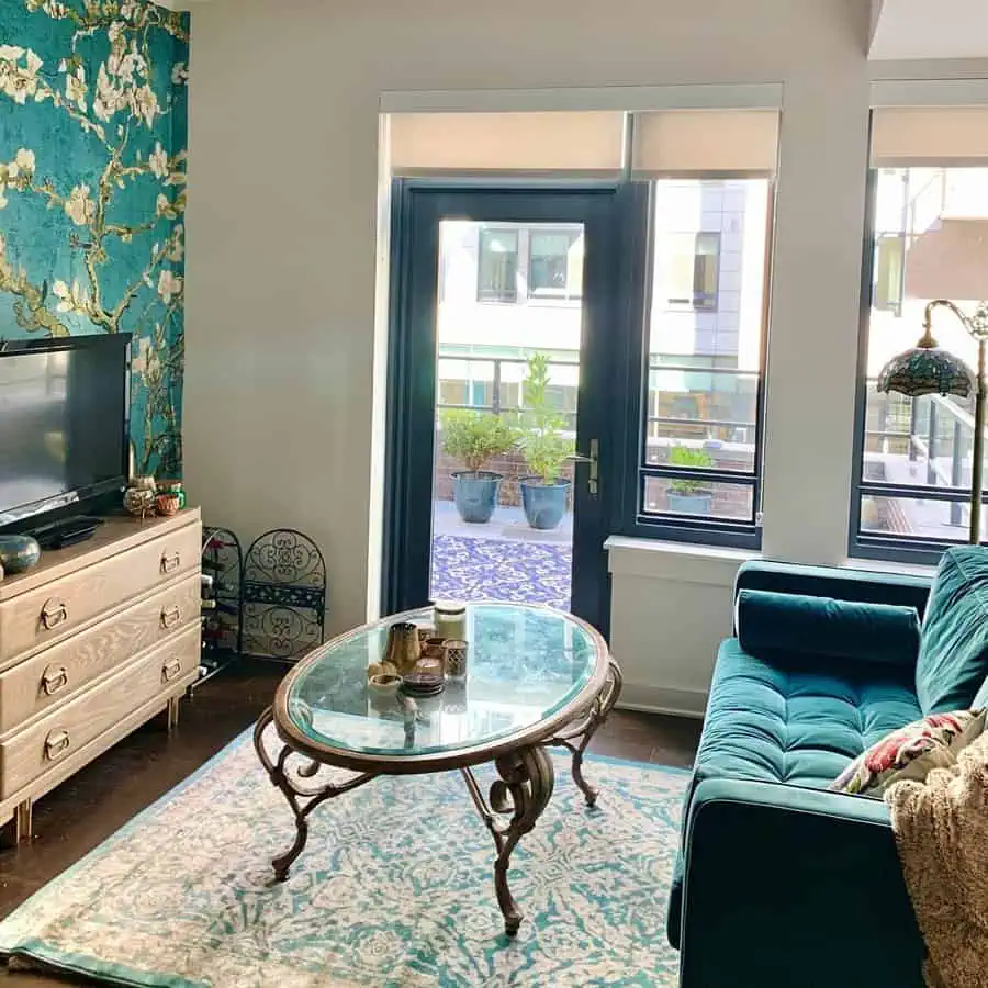 Teal living room with velvet sofa, floral accent wall, glass coffee table, and bright patio access.