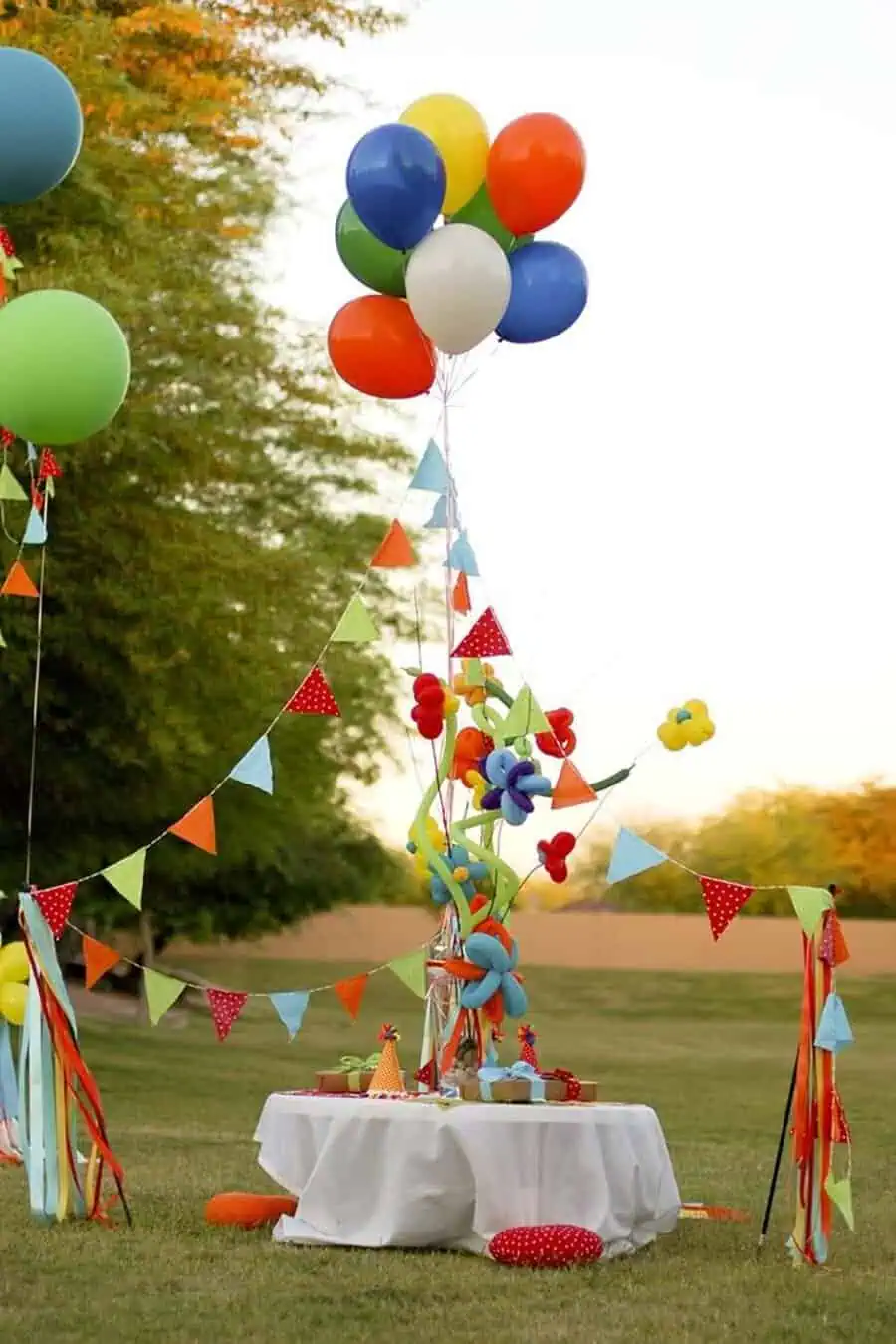 Outdoor birthday table with colorful balloons, bunting, and playful decorations on a grassy lawn.