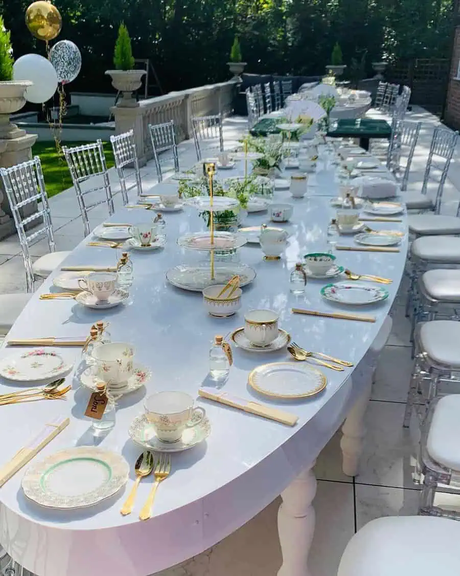 Long outdoor table with vintage tea sets, gold cutlery, and elegant decor for a birthday party.