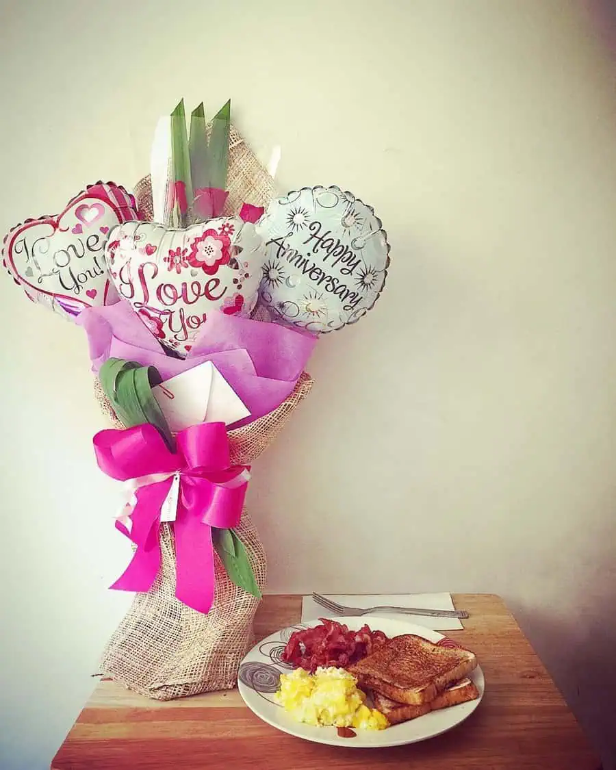 Anniversary bouquet with heart-shaped balloons and a breakfast plate on a wooden table.