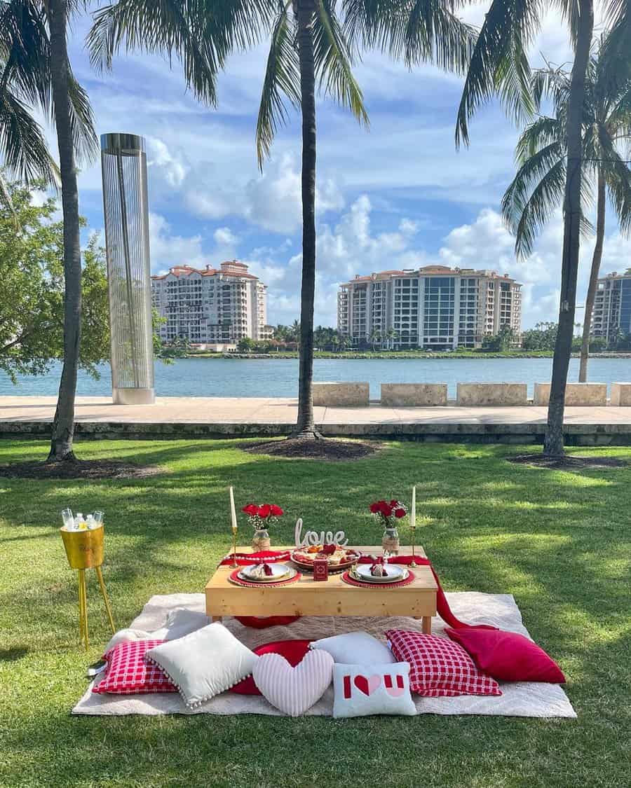 Romantic lakeside anniversary picnic setup with pillows, red roses, and a wooden table.