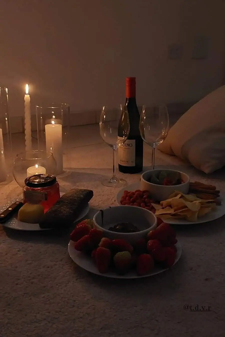 Romantic anniversary indoor picnic setup with wine, candles, strawberries, and light snacks.