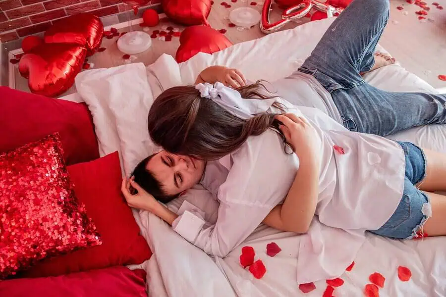 Romantic anniversary setup with a couple on a bed, surrounded by red pillows, balloons, and rose petals.