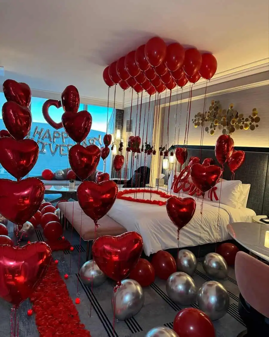 Anniversary room decoration with red heart-shaped balloons, rose petals, and a "Happy Anniversary" sign.