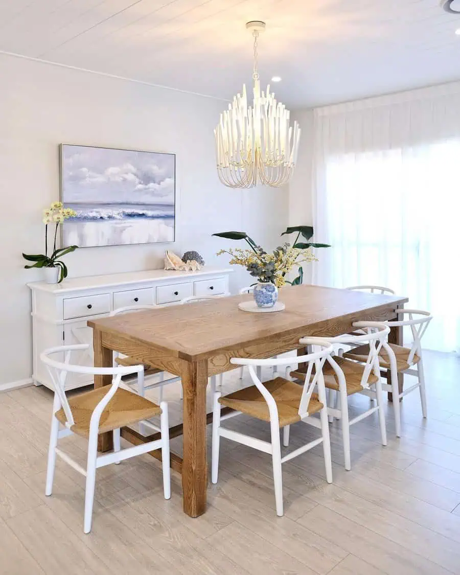 Coastal dining room with a wooden table, white chairs, floral centerpiece, and elegant chandelier.