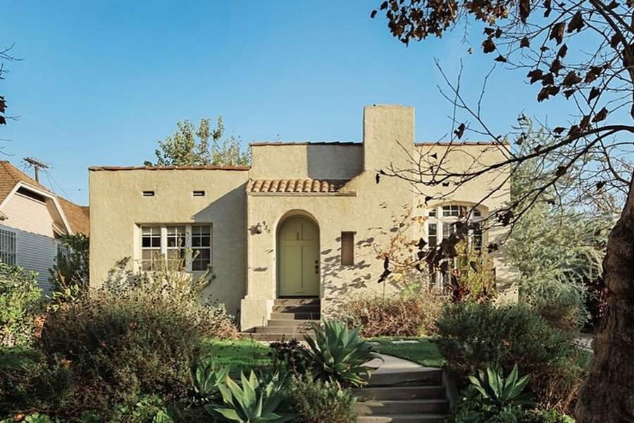 Bungalow-style Spanish home with light beige walls, red tile roof, arched entryway, and lush garden.
