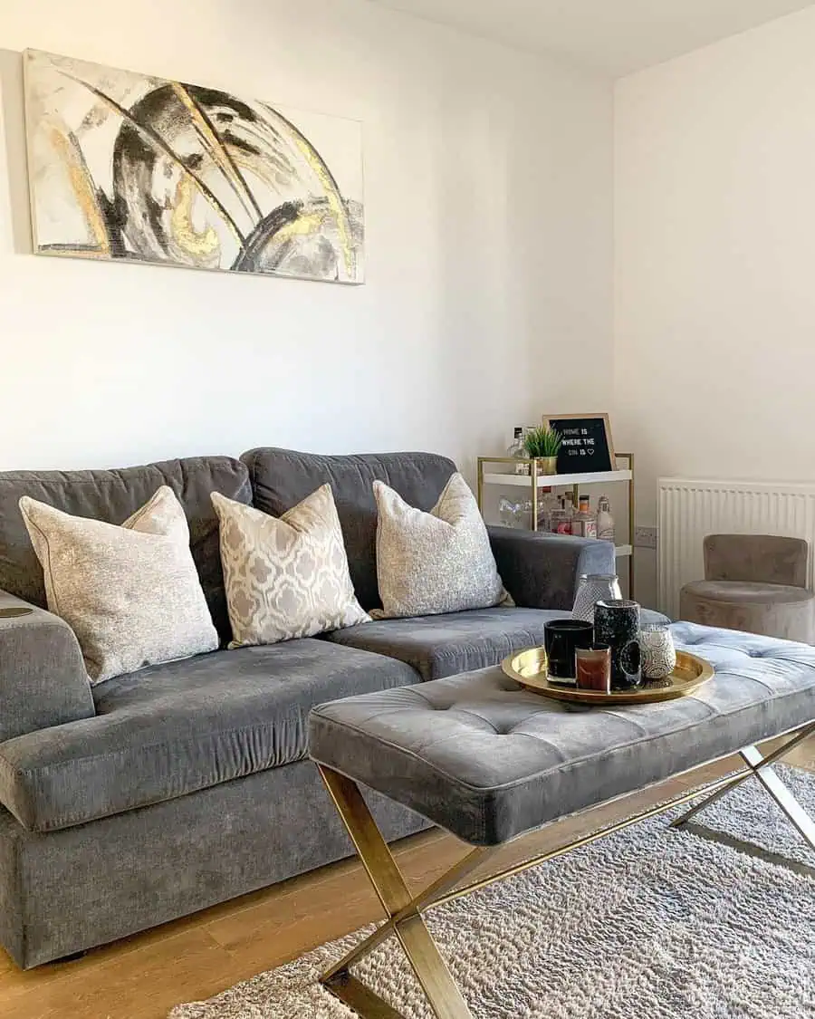 Dark grey sofa with patterned cushions, tufted ottoman, gold accents, and abstract wall art.