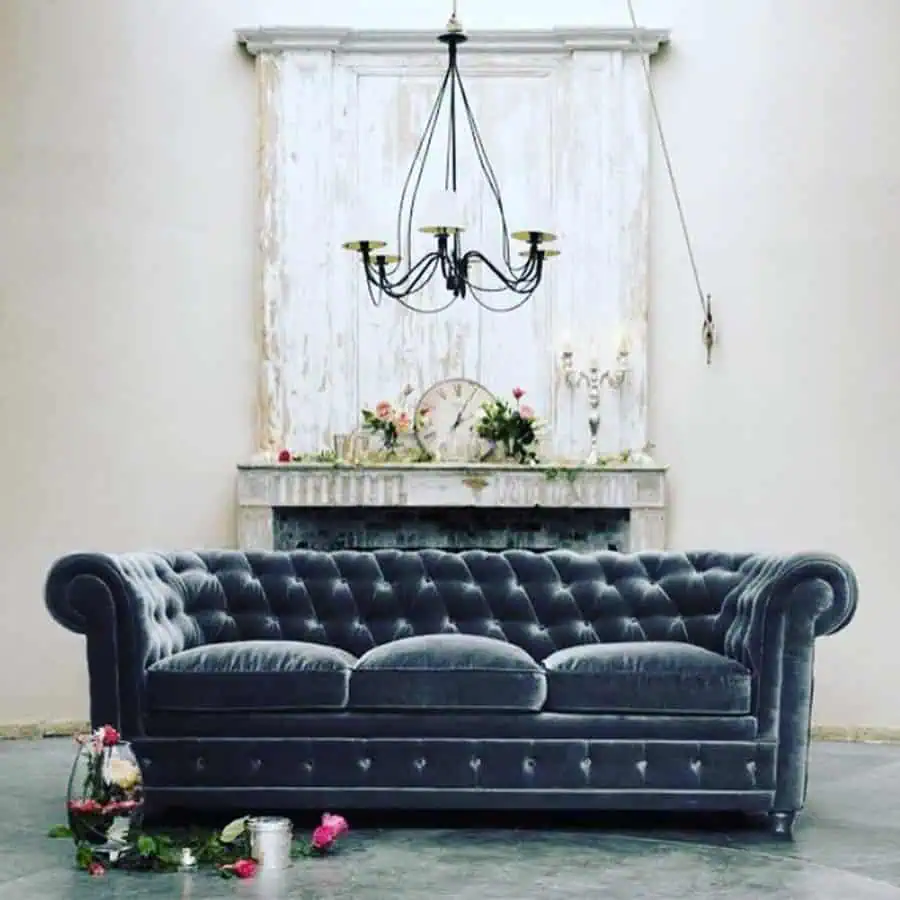 Dark grey tufted velvet sofa with vintage decor, rustic fireplace, and elegant chandelier.