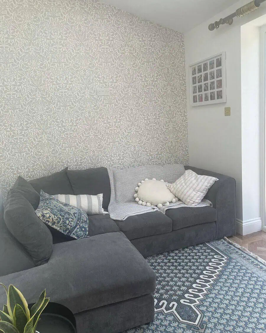 Dark grey sectional sofa with patterned cushions, cozy throws, and floral wallpaper in a bright room.