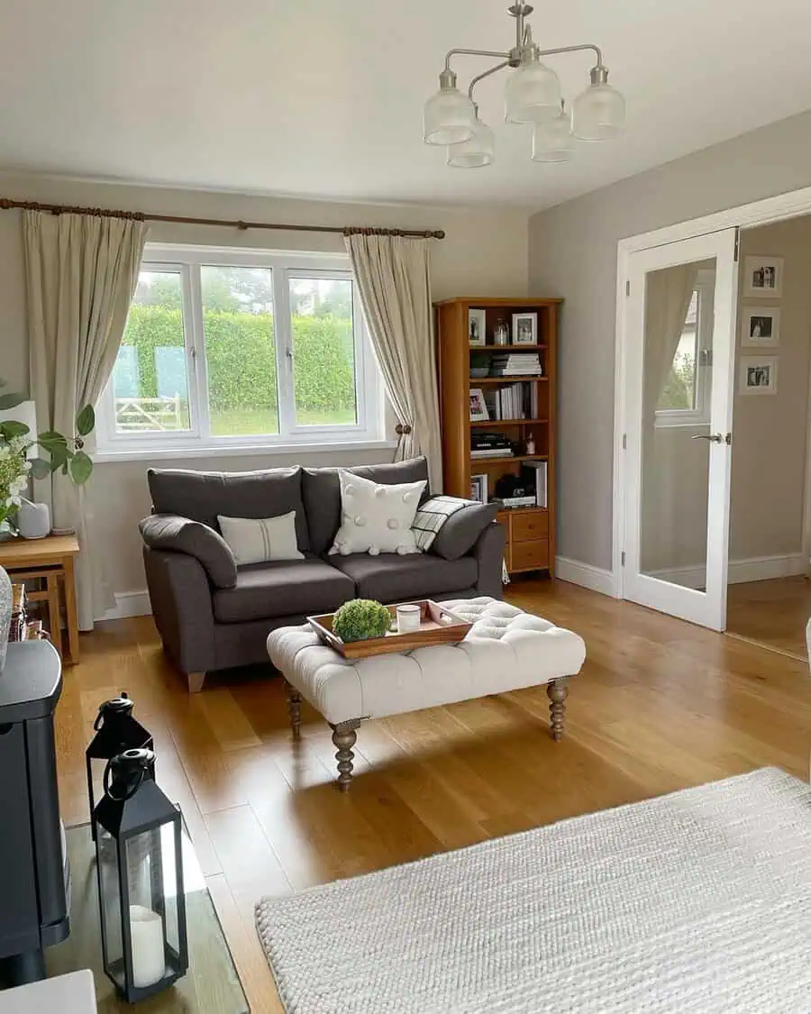 Dark grey sofa with white cushions, light curtains, wooden floors, and a tufted ottoman.