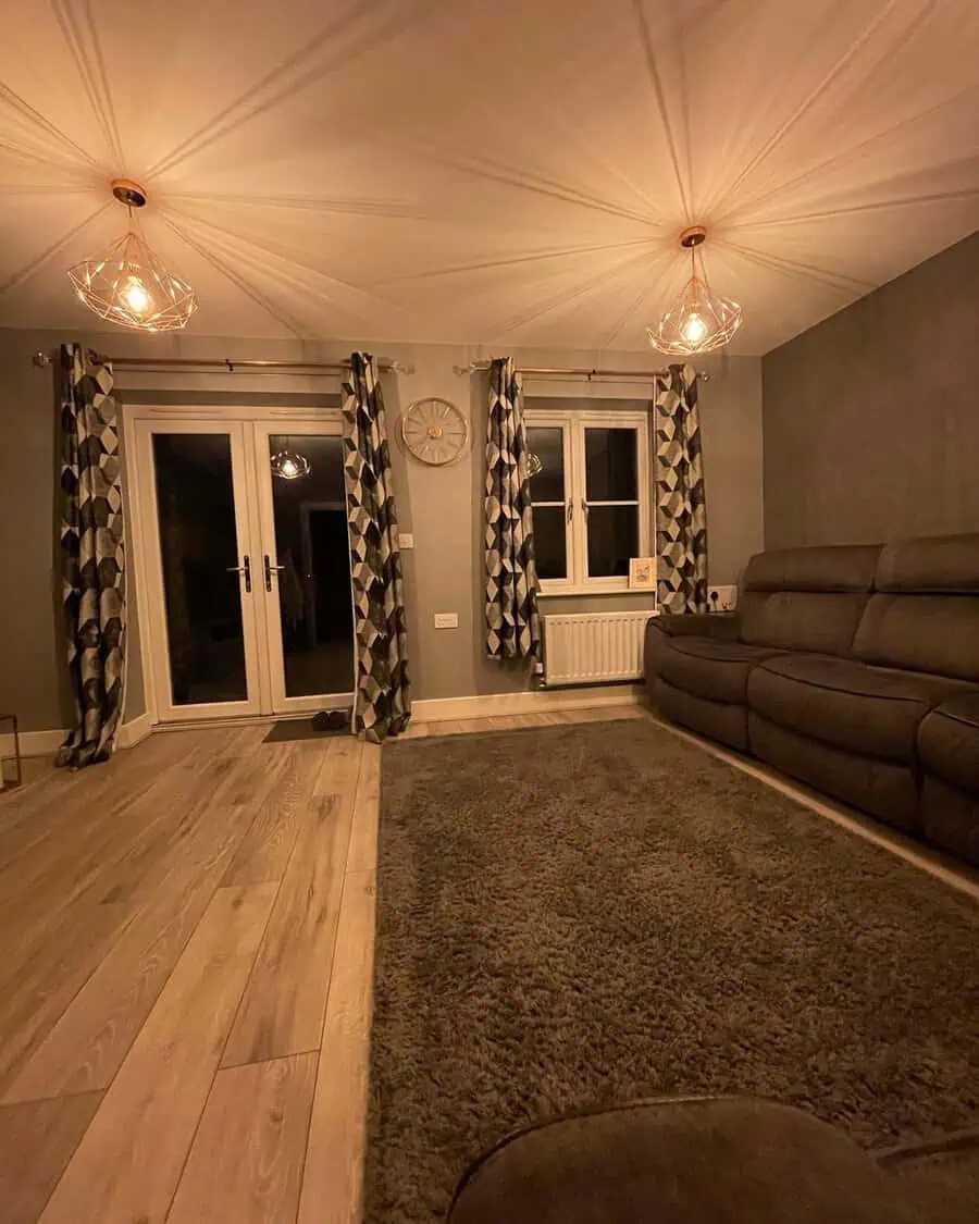 Dark grey reclining sofa with patterned curtains, warm lighting, and a plush rug in a cozy room.