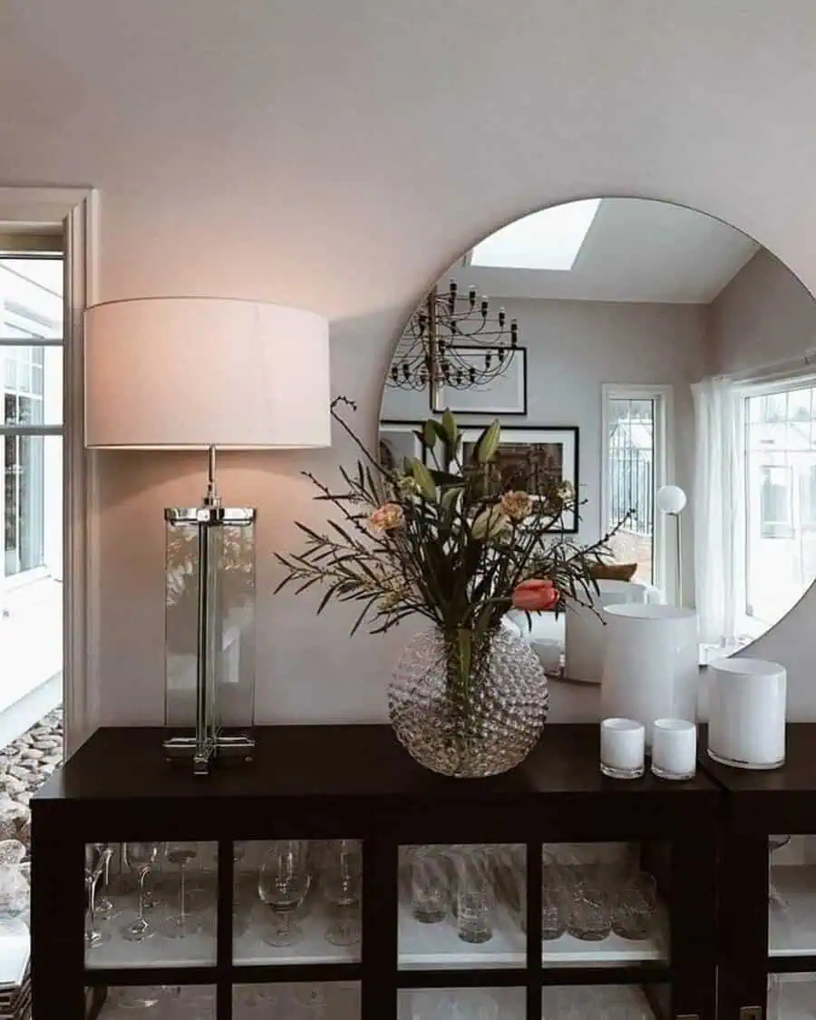 Round mirror on console table with feng shui elements, floral vase, and soft lighting.