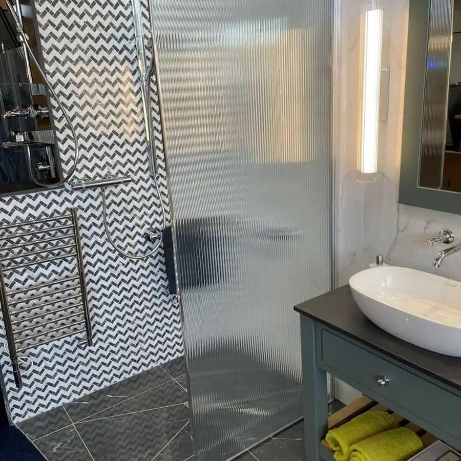 Grey bathroom with herringbone shower wall tiles, fluted glass screen, and black countertop sink.