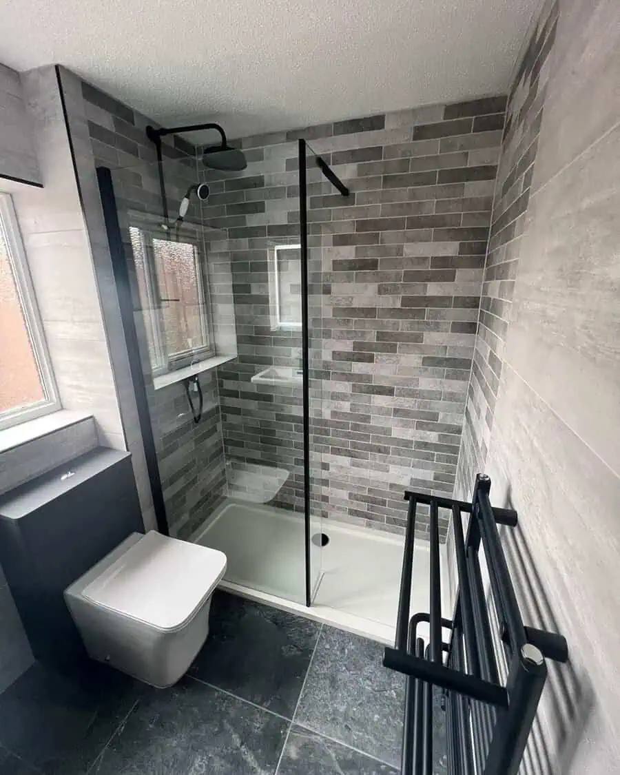 Modern bathroom with grey brick-style wall tiles, dark floor tiles, and glass walk-in shower.