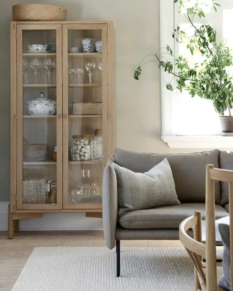 Living room corner with a wooden glass cabinet, cozy sofa, and a large leafy plant near the window.