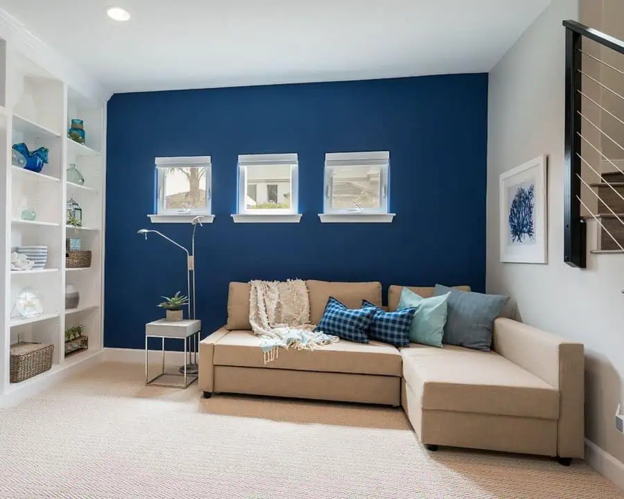 Living room corner with a beige sectional sofa, blue accent wall, built-in shelves, and small windows.