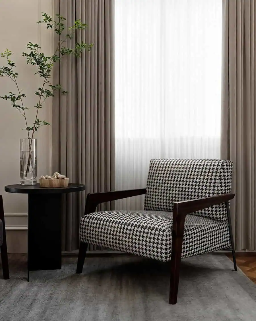 Living room corner with houndstooth armchair, black side table, vase, and neutral curtains.