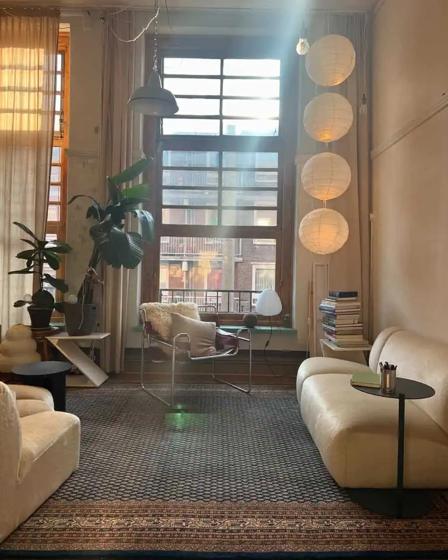 Cozy living room corner with large window, plants, soft seating, paper lanterns, and books.