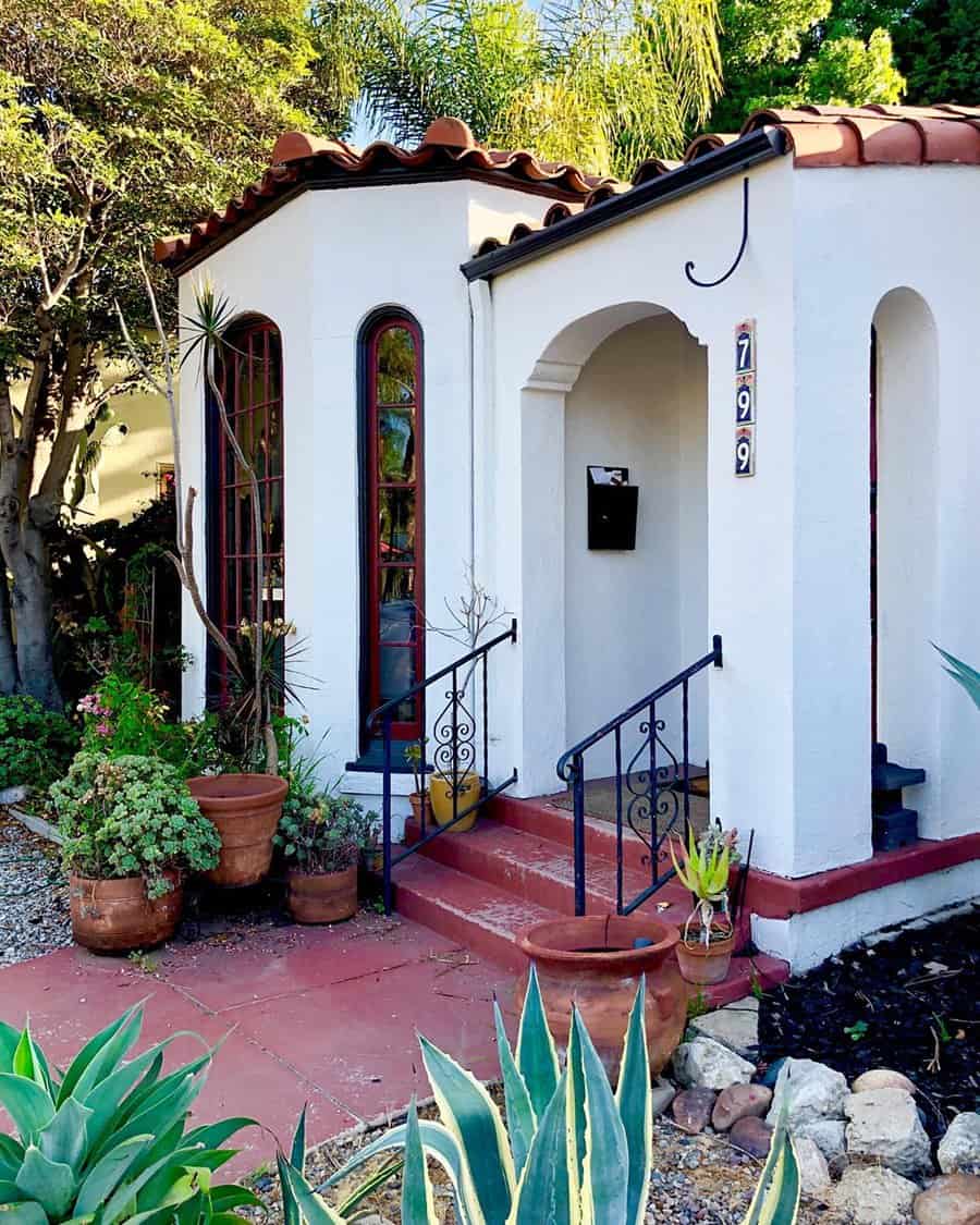 Rustic Spanish-style cottage with white walls, red tile roof, arched entryway, and potted plants.