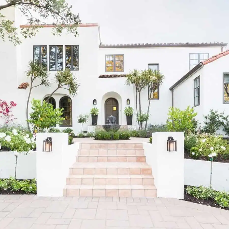 Two-story Spanish-style house with white stucco walls, arched doorway, and landscaped garden.