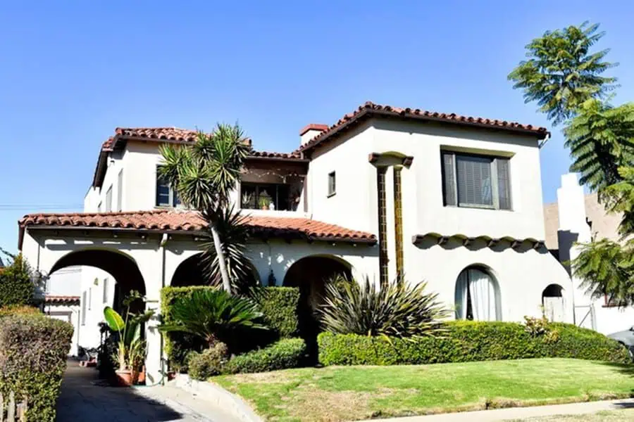 Two-story Spanish-style house with white stucco walls, red tile roof, and lush greenery.