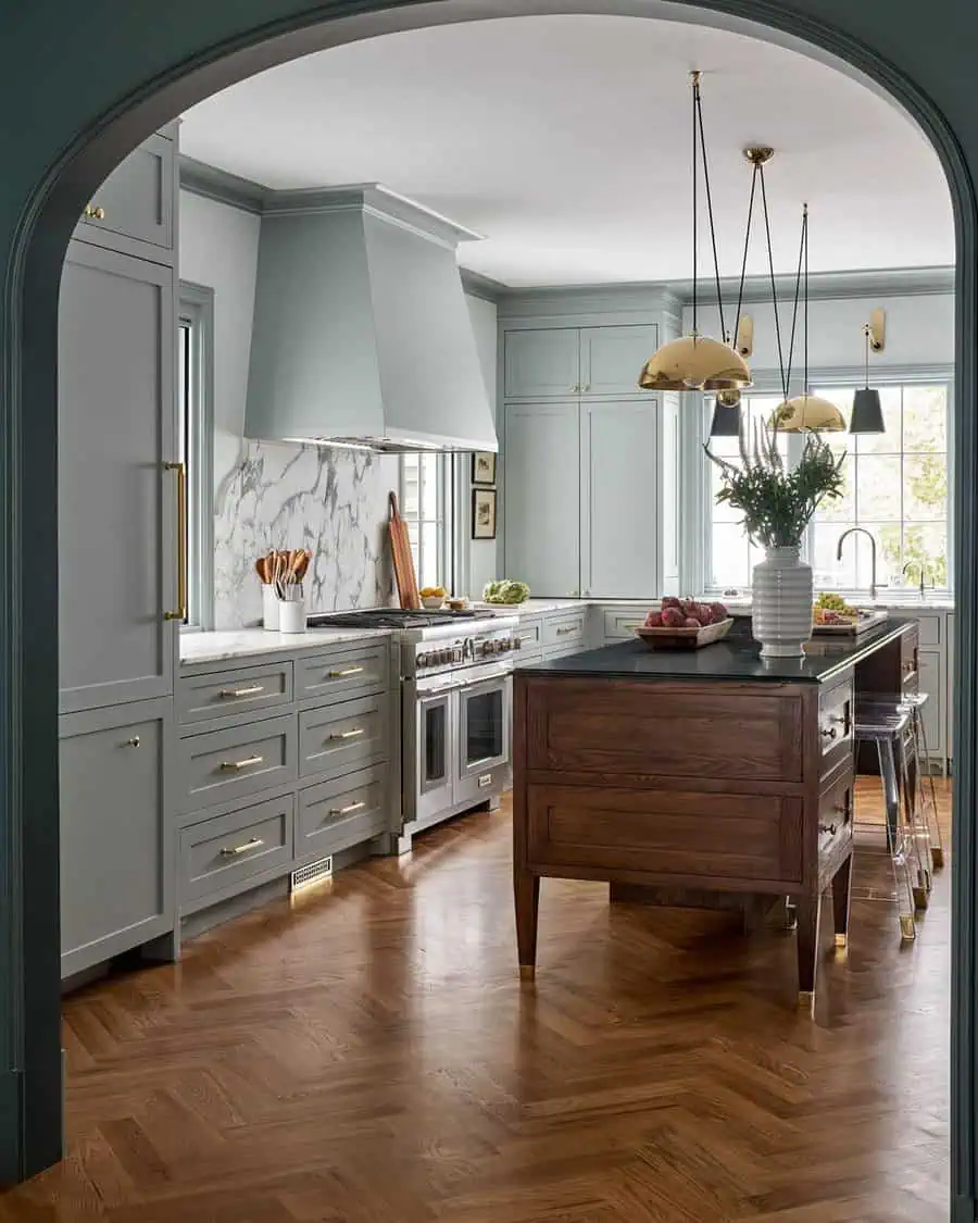 Elegant kitchen with sage green cabinets, marble backsplash, and a wooden island on herringbone floor.