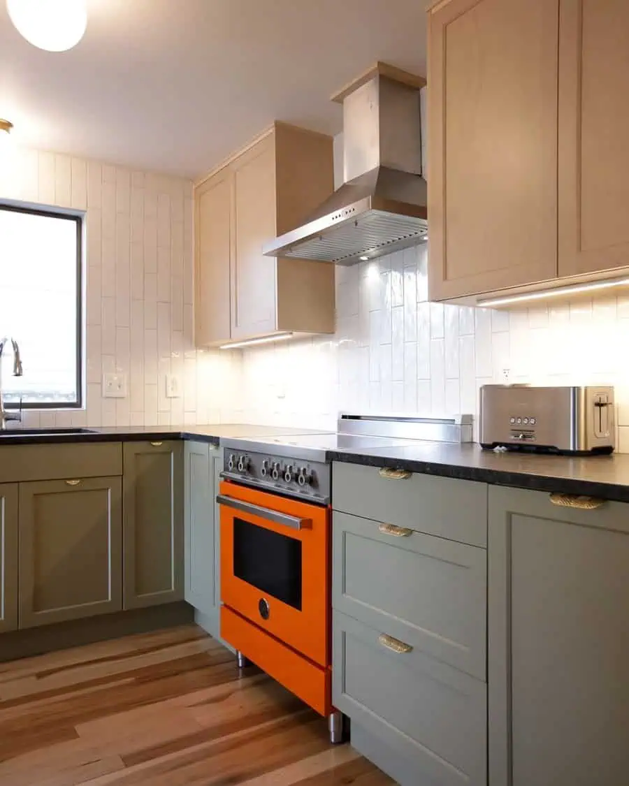 A modern kitchen featuring two-tone cabinets in soft green and beige, highlighted by a bold orange oven and sleek black countertops.