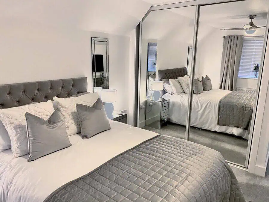 Modern bedroom with a gray and white color scheme, featuring a tufted bed, mirrored wardrobe, and bedside lamps