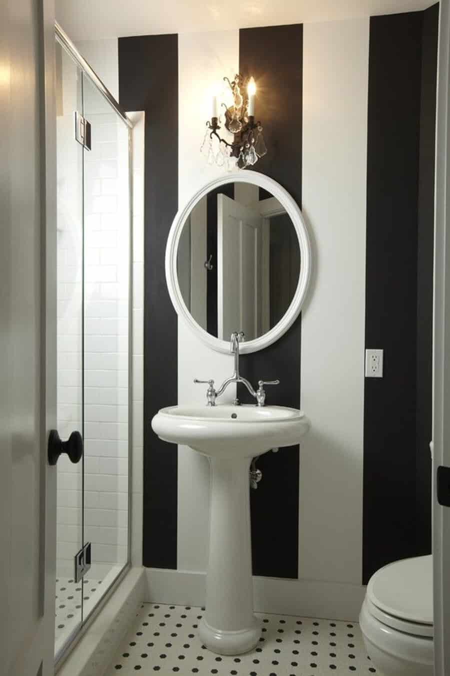 Black and white striped bathroom with pedestal sink, round mirror, and vintage wall sconce.