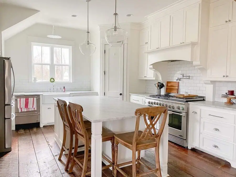 Bright farmhouse kitchen with white cabinets, wood flooring, and a marble island with wooden chairs.