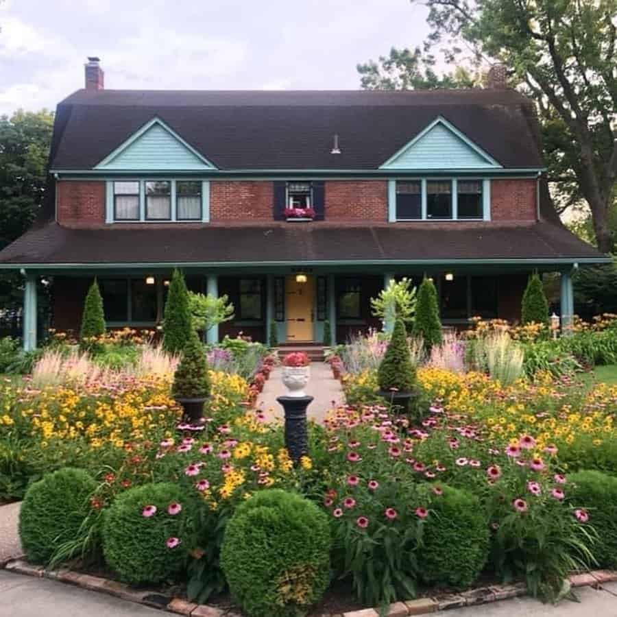 Dutch Colonial house with a wraparound porch, colorful flower garden, and lush landscaping.