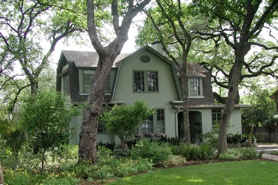 Dutch Colonial house with a stucco finish, gambrel roof, and lush garden surroundings.