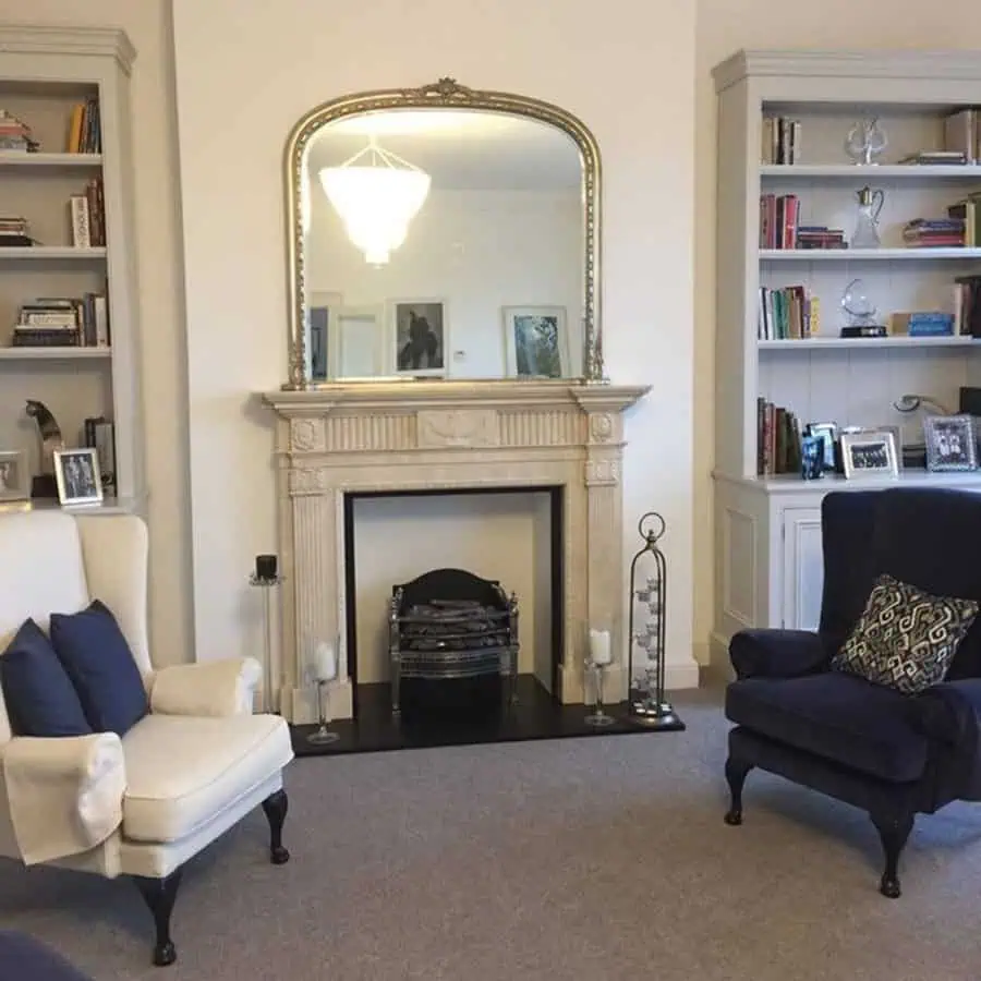 Elegant silver-framed mirror above a marble fireplace with stylish armchairs and bookshelves.