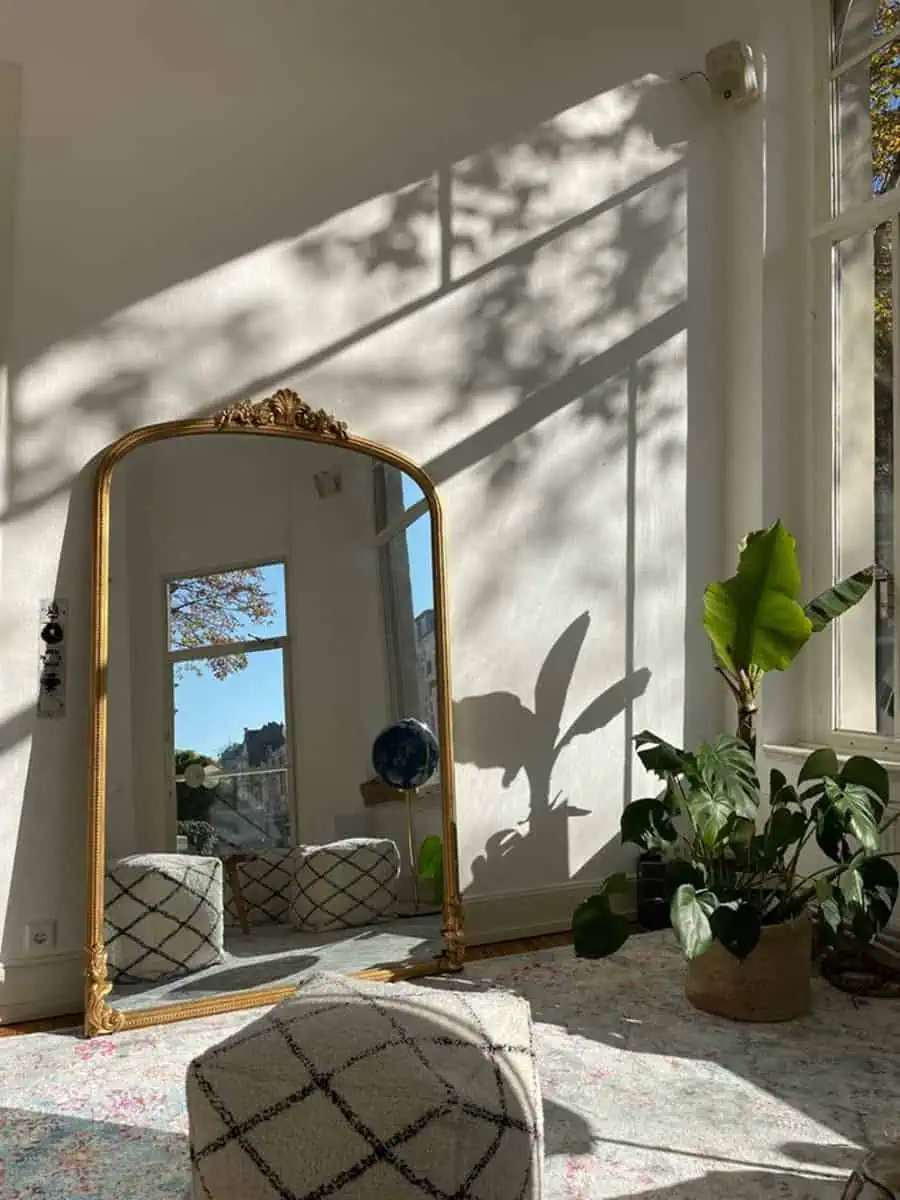 Golden-framed mirror reflecting a cozy space with plants, poufs, and soft lighting.