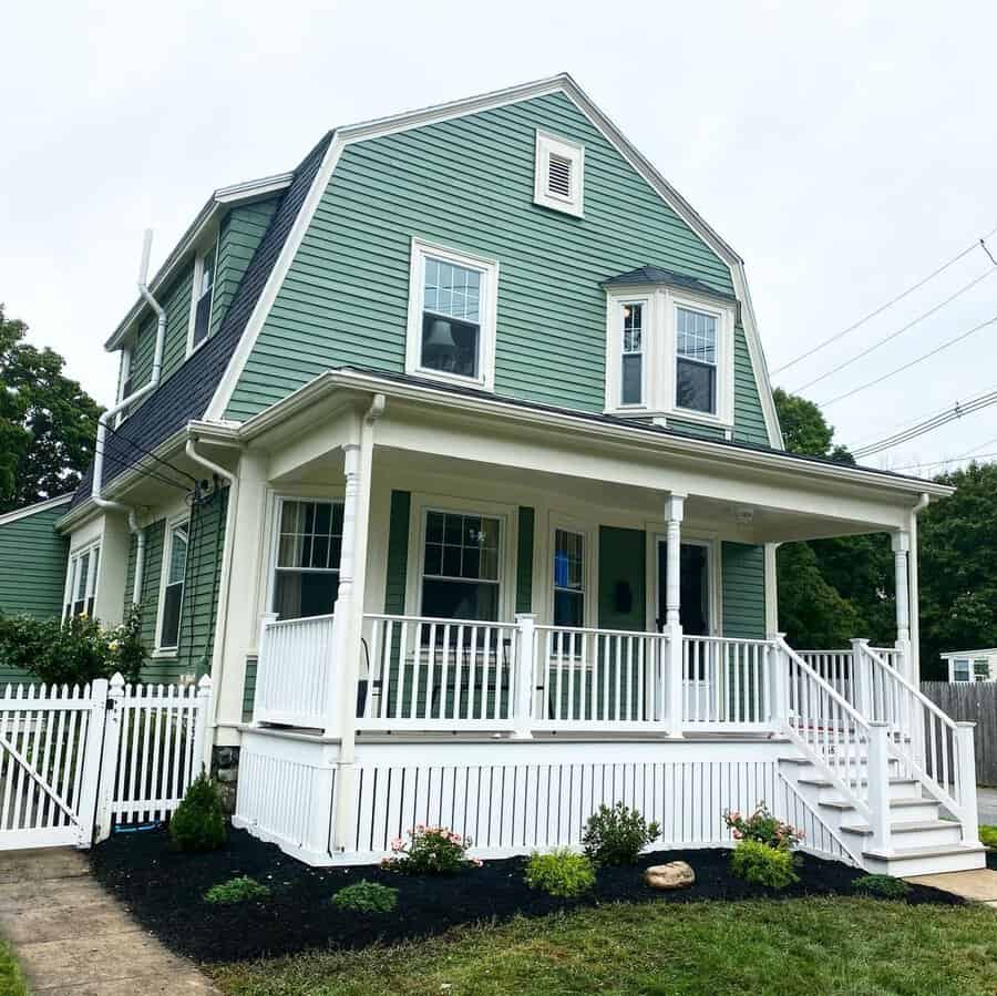 Green Dutch Colonial Revival house with a gambrel roof, wraparound porch, and white trim.