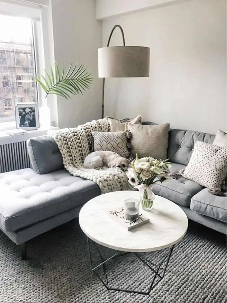 Small grey and white bedroom with a tufted chaise, knit blanket, accent pillows, and a cozy ambiance.