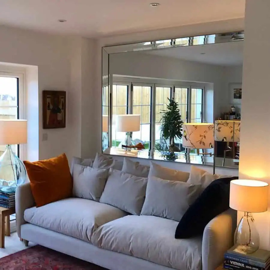 Large frameless mirror reflecting a bright living room with a cozy sofa and stylish decor.