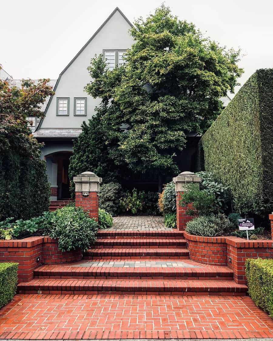 Mid-century Dutch Colonial house with a steep gabled roof, brick pathway, and lush greenery.