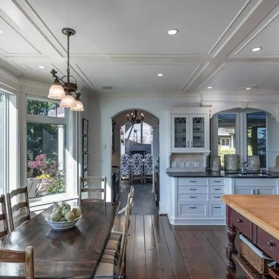 Bright kitchen with a modern coffered ceiling, wooden dining table, white cabinetry, and large windows.