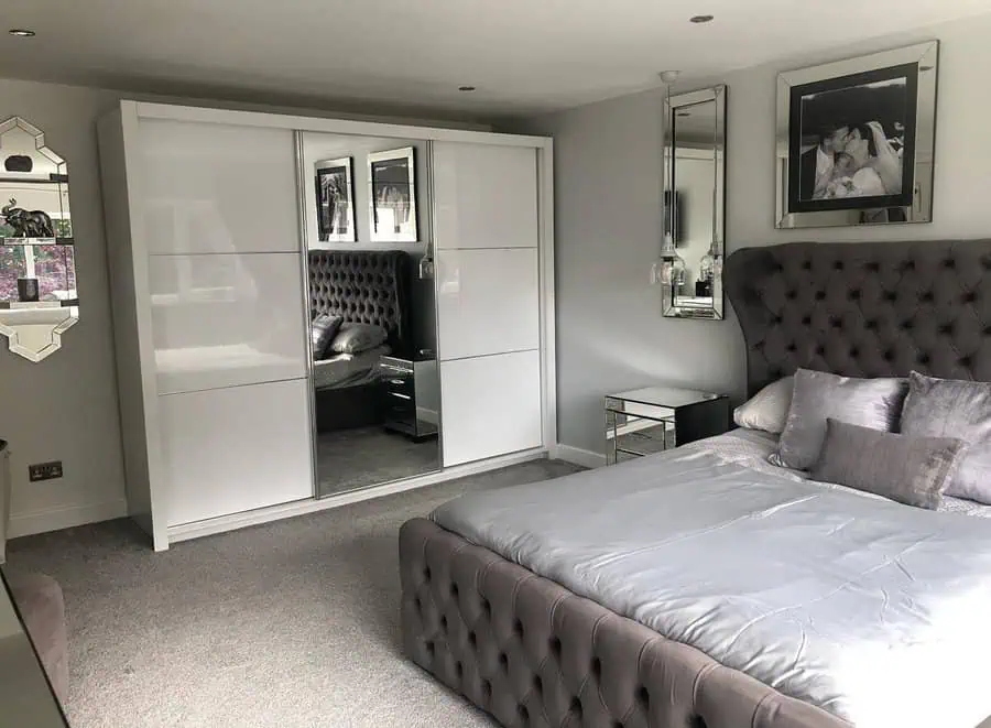 Luxurious grey bedroom with a tufted bed, mirrored furniture, and sleek white wardrobe.