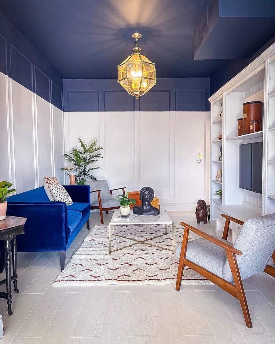 Navy blue living room with white panel walls, a velvet sofa, gold lighting, and mid-century chairs.