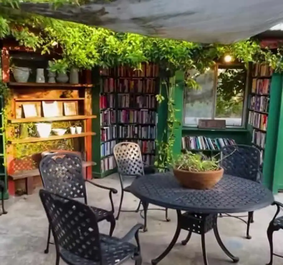 Outdoor home library with lush greenery, metal patio furniture, and bookshelves under a canopy.