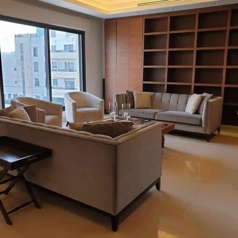Modern living room with built-in wooden bookshelves, neutral-toned furniture, and large windows.