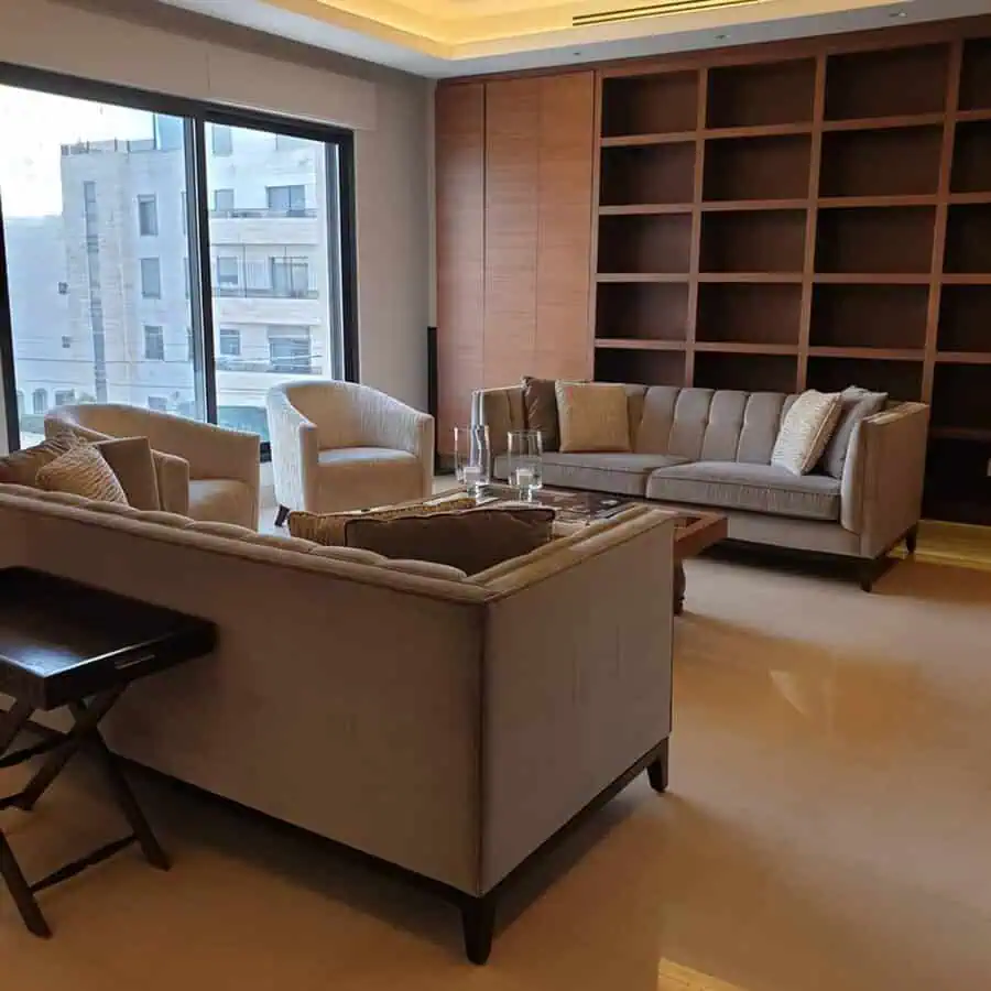 Modern living room with built-in wooden bookshelves, neutral-toned furniture, and large windows.