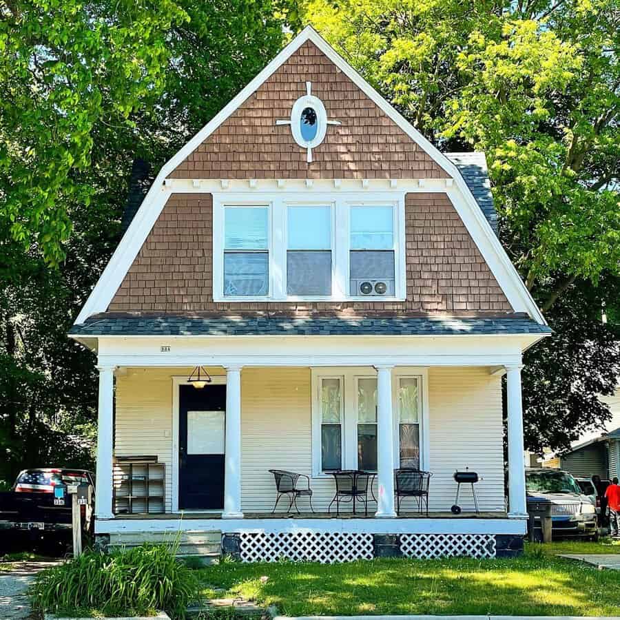 Two-tone Dutch Colonial house with brown shingle siding, white trim, and a covered front porch.