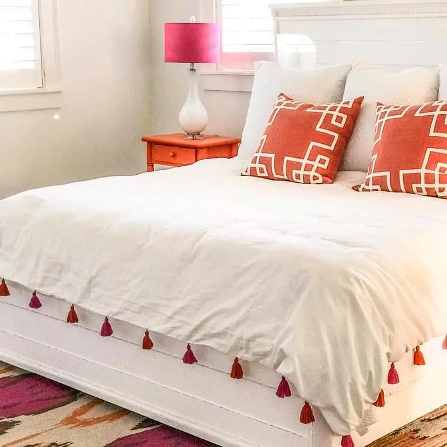 White bedding with orange accent pillows, a tassel-trimmed duvet, and a bold nightstand for contrast.